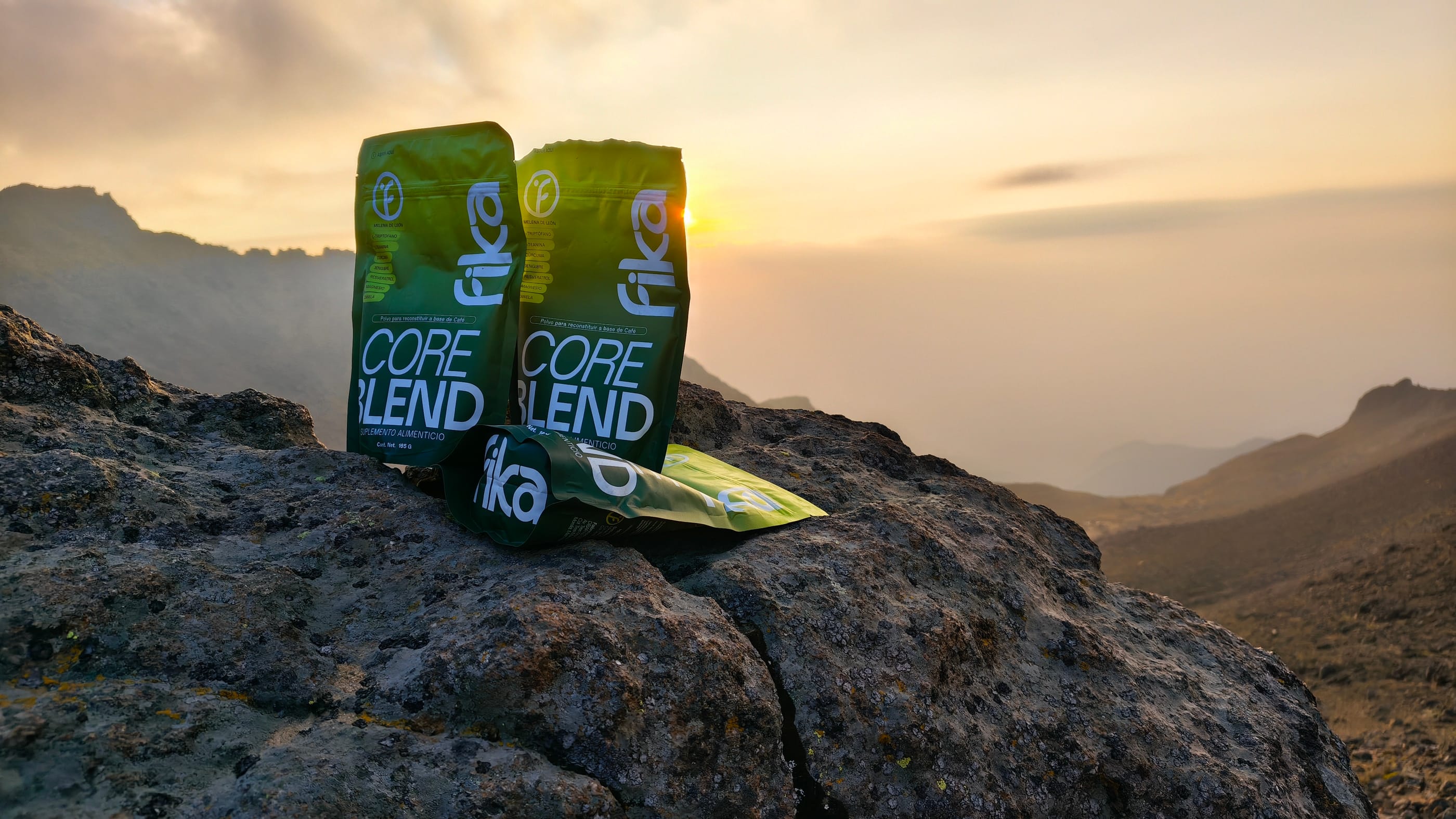Tres bolsas de FIKA Core Blend sobre una roca, con el sol saliendo entre las montañas